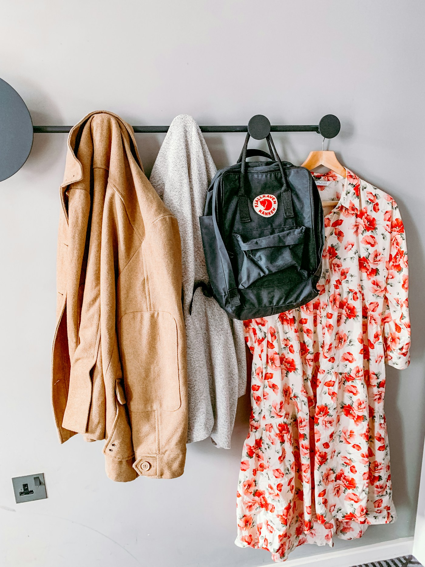 Coats, a backpack and a floral dress hanging from a wall-mounted rail on a white wall.