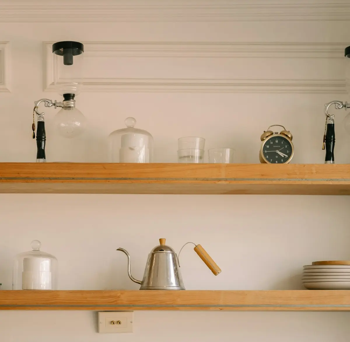 Open wooden kitchen shelves arranged with white ceramics, a metal kettle, glasses and a small clock